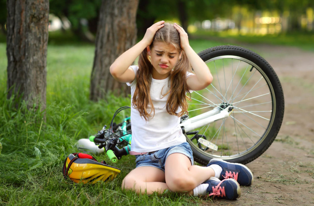 A young girl on a bicycle fell to the ground, sitting and holding her head with both hands.