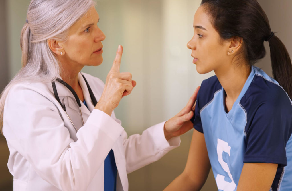 A lady doctor in a white lab coat is standing in front of a young girl wearing an athlete uniform that sitting face to face. The doctor's index finger pointing upward in front of the patient seems like assessing the patient eye responsiveness.