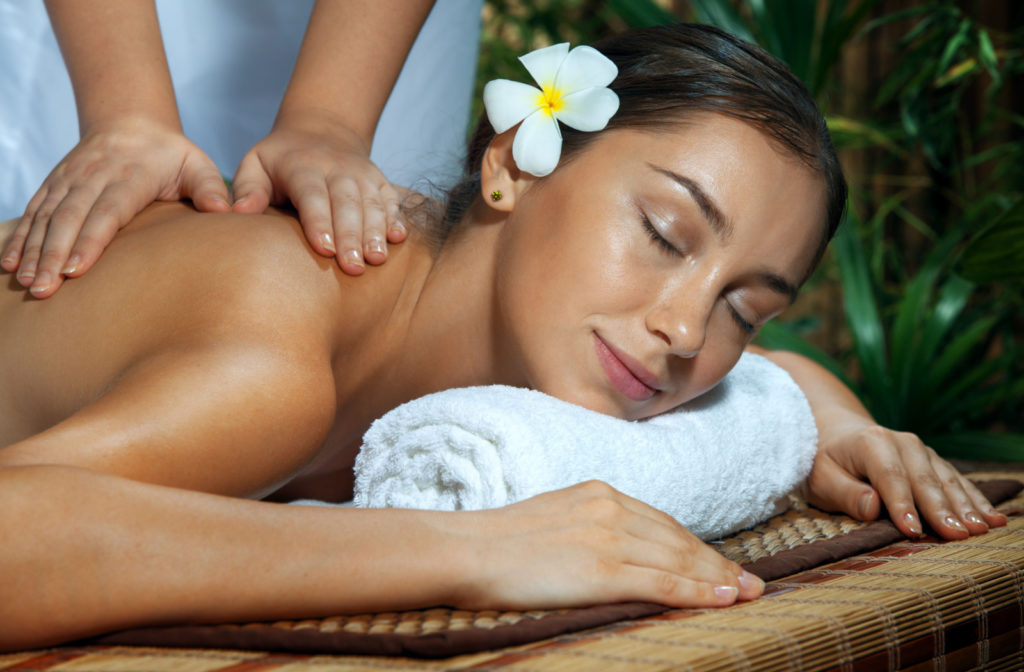 A close-up of a young woman with a white flower in her hair looking relaxed while getting massage therapy
