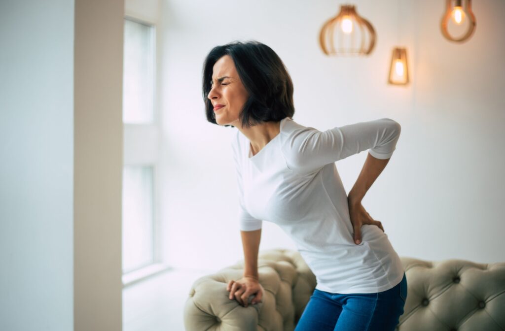 A woman with one hand on the couch for support and the other on her lower back as she experiences chronic pain while standing up.