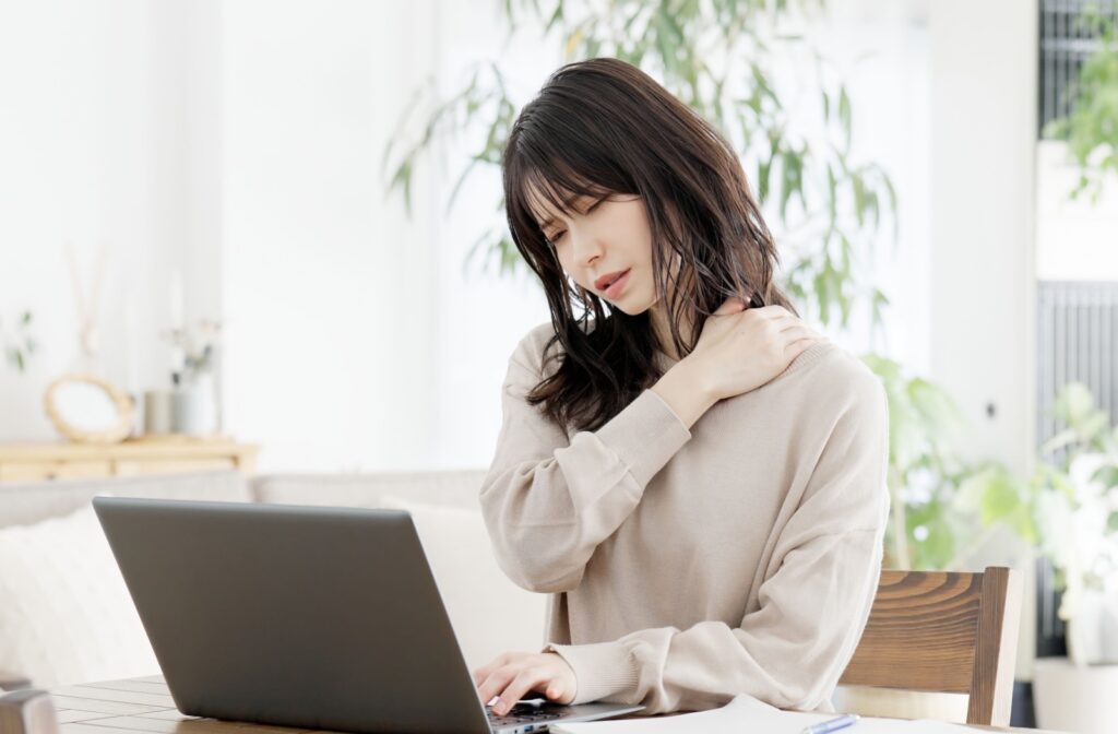 A young adult at home working on their laptop rubbing their neck in discomfort due to pain from a whiplash injury.