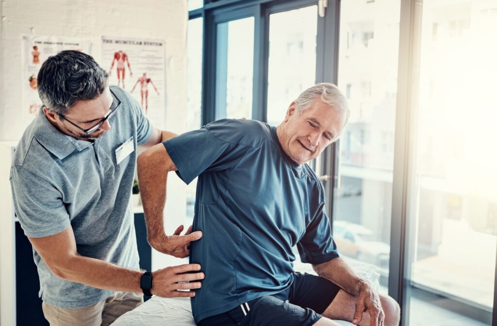 An older adult gesturing to their lower back while a physiotherapist examines the area.