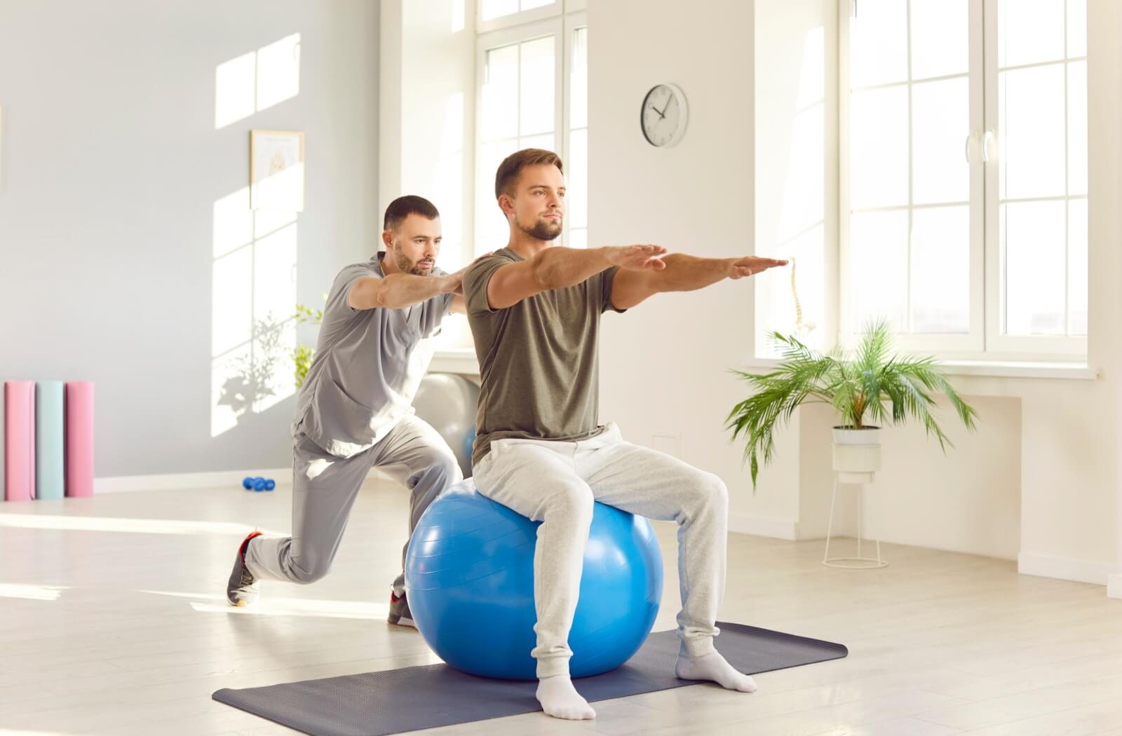 A person sits in comfortable clothing on a balance ball during an exercise with their physical therapist.